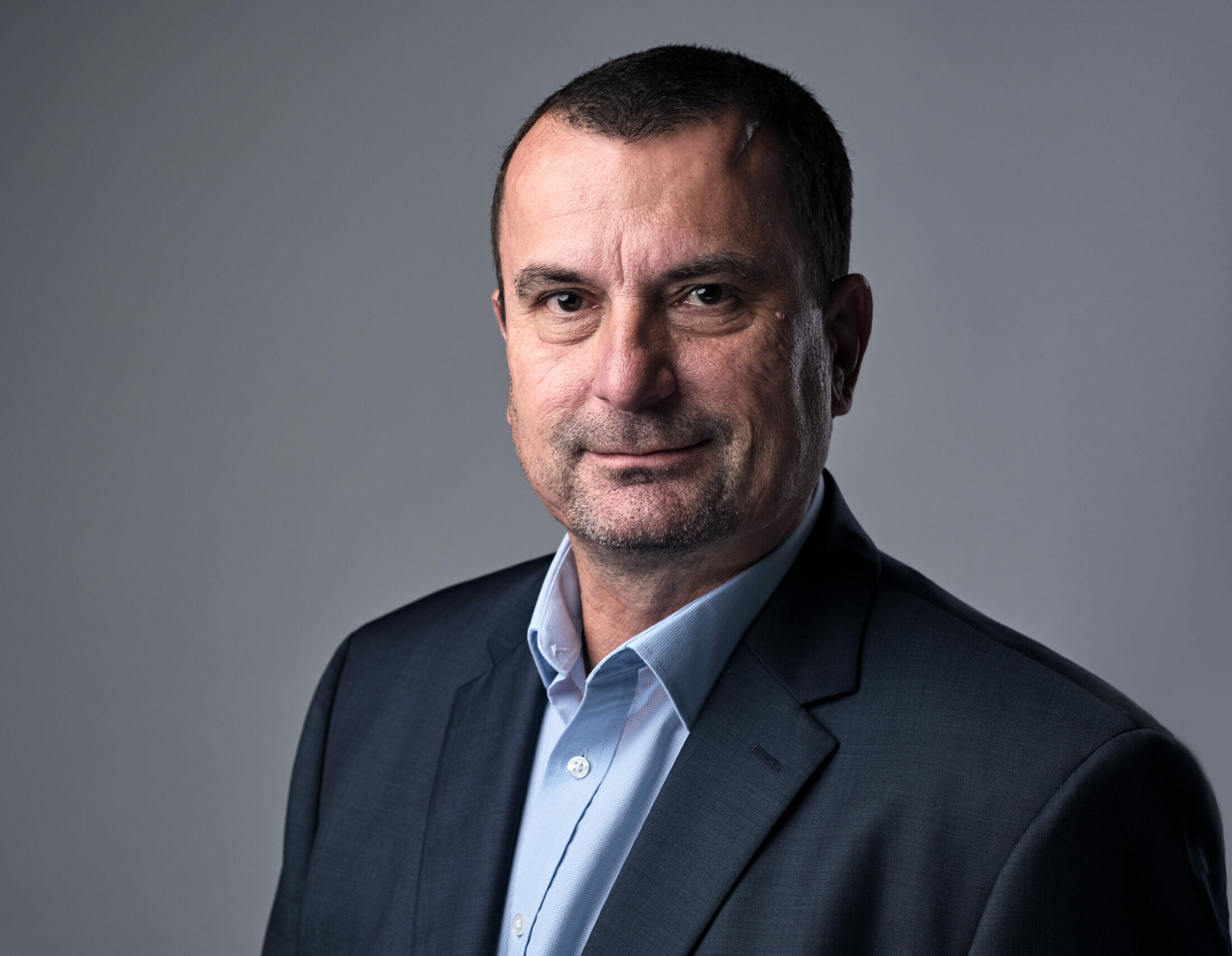 middle aged man with short dark hair looking serious in a blue shirt and dark jacket