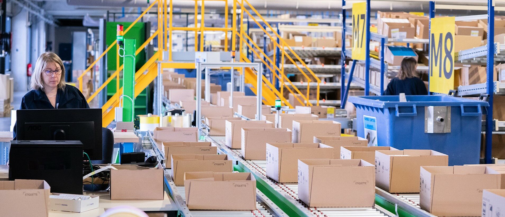 Woman staff focusing on computer screen in warehouse with packages on conveyor belt